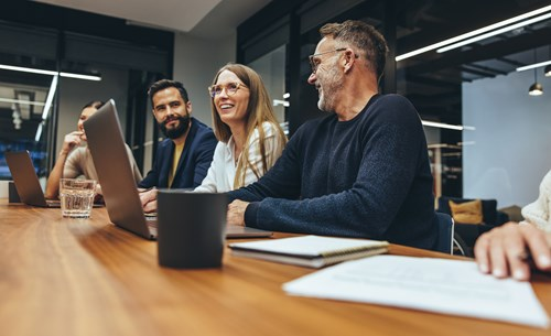 A group of people sitting at a conference table with laptops, engaged in a discussion inside a modern office space.