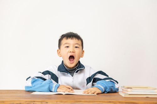 A young boy in a striped jacket yawning at a wooden table with books next to him, against a plain white background.