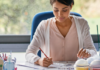 Student-focused assessment A person sitting at a desk drawing on a large sheet of paper with coloured pencils, surrounded by art supplies, in a bright room with a large window.