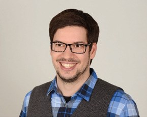 A man wearing glasses, a blue plaid shirt, and a grey vest smiling at the camera against a plain background.