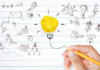 Grammar me this, grammar me that A hand holding a pencil drawing a concept illustration of a light bulb with a crumpled yellow paper as the bulb, surrounded by various sketched symbols like graphs, gears, question marks, and other ideas on lined paper.