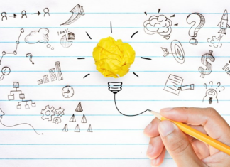 Grammar me this, grammar me that A hand holding a pencil drawing a concept illustration of a light bulb with a crumpled yellow paper as the bulb, surrounded by various sketched symbols like graphs, gears, question marks, and other ideas on lined paper.