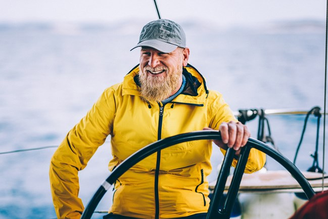 A bearded man wearing a yellow rain jacket and a cap is smiling while steering a sailboat.