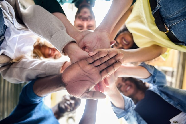 A diverse group of people standing in a circle with their hands stacked in the center, photographed from below.