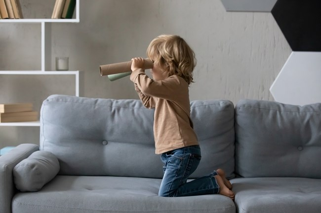 A young child kneeling on a grey sofa, looking through a homemade telescope made from paper tubes, in a modern living room setting.