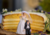 Value or valued? Are you dating or married? A partially sliced layered cake with a humorous wedding cake topper featuring a bride standing with her hand on her hip and a groom kneeling beside her, set against a wooden base and decorated with small flowers.