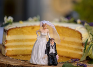 Value or valued? Are you dating or married? A partially sliced layered cake with a humorous wedding cake topper featuring a bride standing with her hand on her hip and a groom kneeling beside her, set against a wooden base and decorated with small flowers.