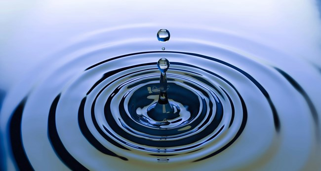 A single water droplet falling into a calm body of water, creating concentric ripples that spread outward.