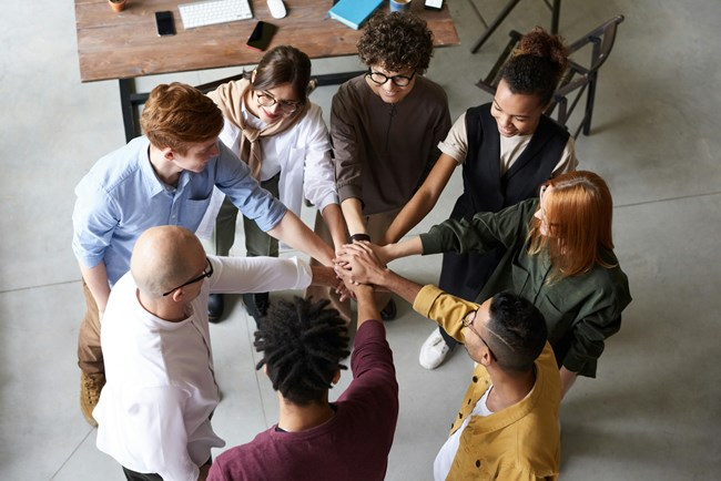 A diverse group of eight people standing in a circle with their hands joined in the center, symbolizing unity and teamwork. They are in a casual office setting with a wooden desk and office supplies in the background.
