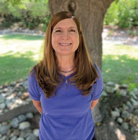 A woman with long brown hair and a purple shirt standing in front of a tree, smiling at the camera.