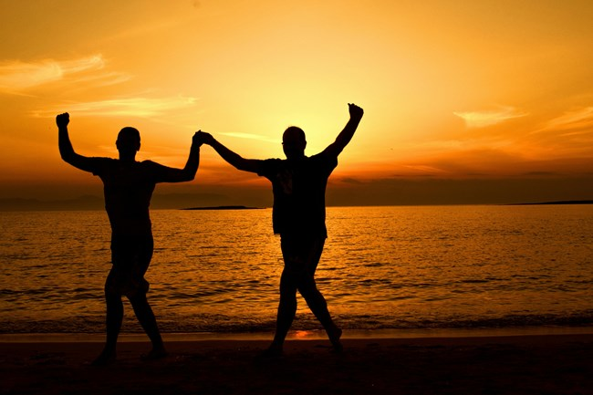 Two people holding hands in silhouette against a vibrant orange sunset over the sea, walking along a beach.