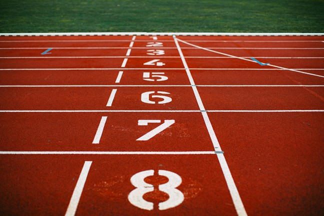 A view of a red running track with eight numbered lanes, leading to a grassy field in the background.
