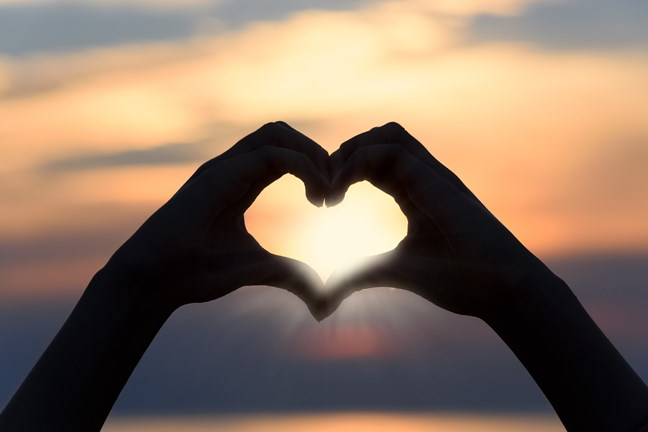 Silhouette of hands forming a heart shape with the sun shining through against a sunset background.
