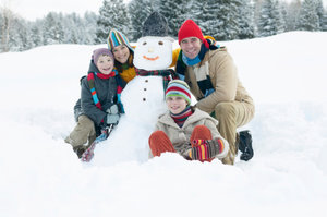 A family of four, wearing colorful winter clothing, poses with a snowman in a snowy landscape with trees in the background.