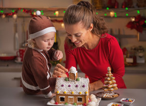 A woman and a child in festive clothing are decorating a gingerbread house at a table, surrounded by Christmas decorations and lights.