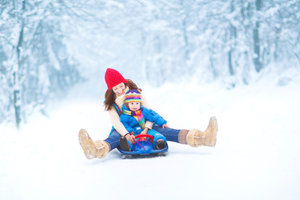 A woman and a child wearing colorful winter clothing slide down a snowy path on a sled surrounded by snow-covered trees.