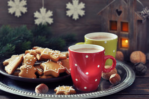 Two polka dot mugs filled with hot beverages sit on a silver tray beside a plate of star and tree-shaped cookies, with a wooden lantern and snowflake decorations in the background.