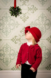 A young child dressed in a red cardigan and red hat looks up at a mistletoe hanging from the ceiling, against a vintage patterned wallpaper background.