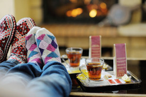 Two people with cosy patterned socks relaxing in front of a fireplace with two trays of tea and biscuits on a table.