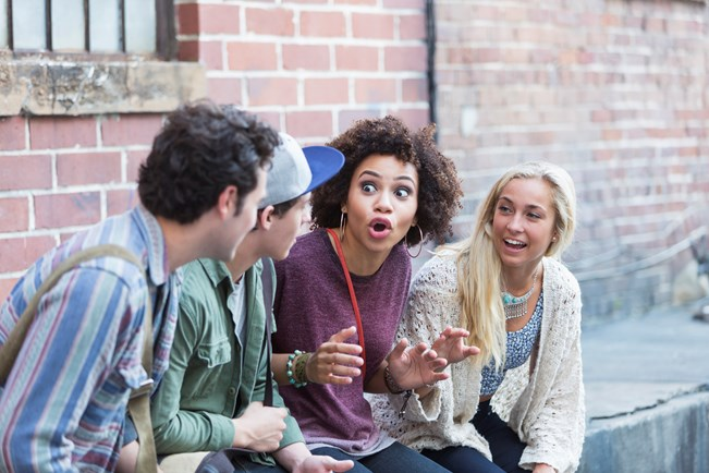 A group of four young adults sitting outside, engaged in animated conversation; the person in the middle is gesturing expressively, while the others listen attentively and smile.
