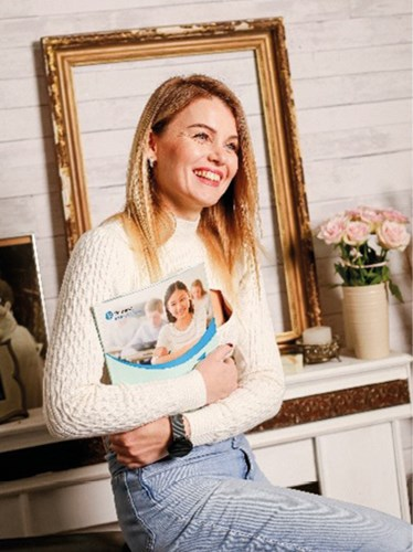A woman with long blonde hair, wearing a white sweater and blue jeans, is smiling while holding a brochure. She is sitting in a cozy room with a framed picture and a vase of pink flowers on a mantel behind her.