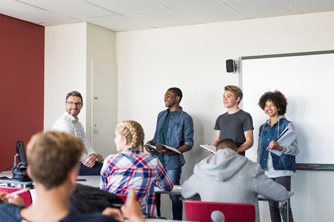 Four students standing at the front of a classroom with a teacher, facing a seated audience.
