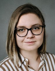 A person with shoulder-length straight hair, wearing glasses and a striped shirt, poses against a neutral background.