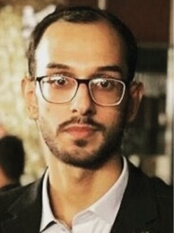 A man with glasses and a beard wearing a suit, gazing directly at the camera in a blurred indoor setting.