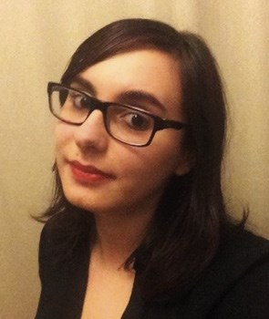 A person with dark hair wearing glasses and a black top, posing against a beige background.