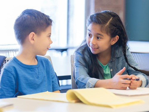Two young children sitting at a table, smiling and talking to each other, with papers in front of them in a classroom setting.