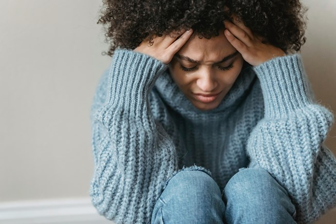 A woman sitting with a distressed expression, holding her head with both hands, wearing a knitted blue sweater and jeans.