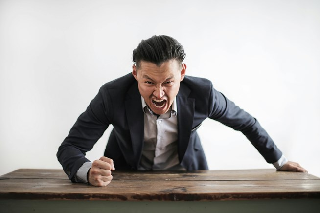 A man in a suit angrily leans over a desk, clenching his fist and shouting.