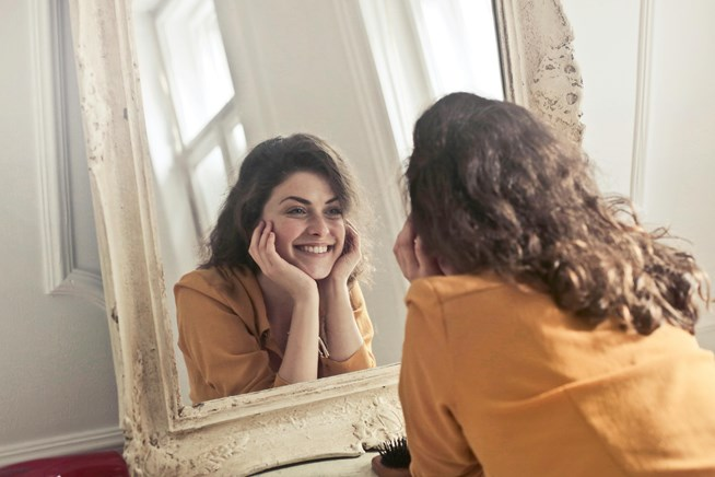 A woman in an orange top smiles while looking at herself in an antique-style mirror, her chin resting on her hands.