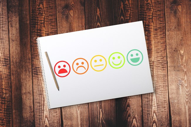 A notebook with a series of six coloured emoticons ranging from sad to happy on a wooden table, with a pencil placed beside it.