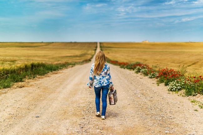A woman with long hair wearing a blue floral blouse and jeans walks down a long dirt road through a rural landscape, carrying a suitcase, with fields and wildflowers on either side under a clear sky.