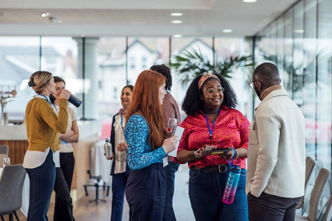 A group of people socializing in a modern office setting; one woman holds a tablet and engages in conversation.
