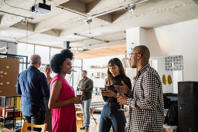 A group of people socializing at an indoor gathering, with one woman in a pink dress holding a drink, another woman looking at her phone, and a man in a checked shirt talking to them. The room has a modern, industrial style with exposed ceilings and large windows.