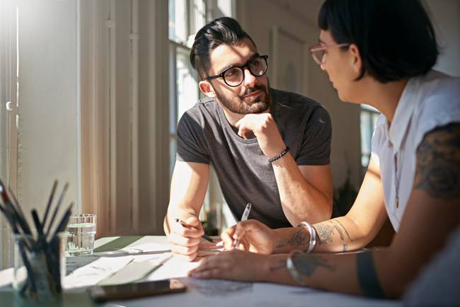 Two people sit at a table in a sunlit room, engaged in a discussion with papers and pens in front of them.