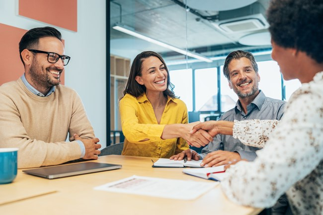 Four people sitting around a conference table, two of them smiling and shaking hands, while the other two look on with smiles, creating a positive business meeting atmosphere.