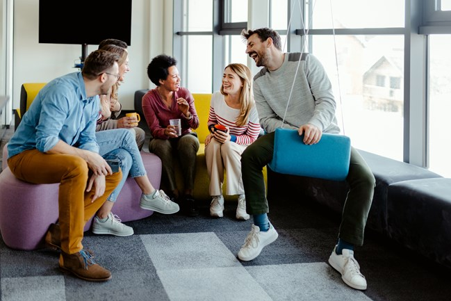 A group of six people sitting and standing together in a modern office setting, smiling and holding cups as they engage in a casual conversation near large windows.