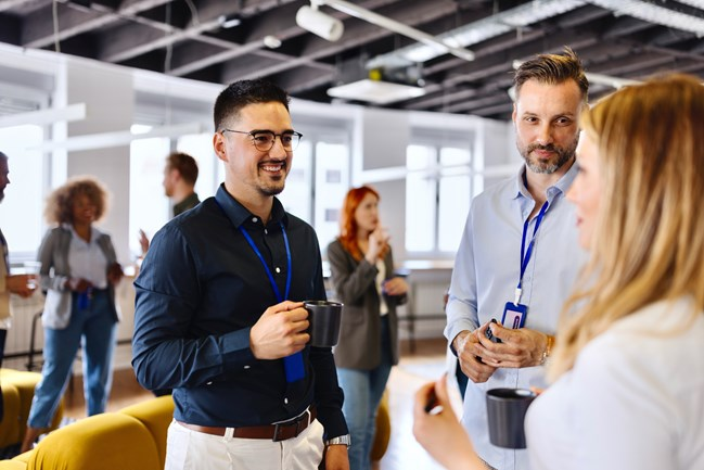 A group of professionals in an office setting, conversing and holding coffee mugs, wearing blue lanyards.