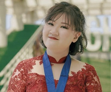 A person smiling, wearing a red lace dress and a blue ribbon around their neck, standing outside in a sunlit area with blurred greenery in the background.
