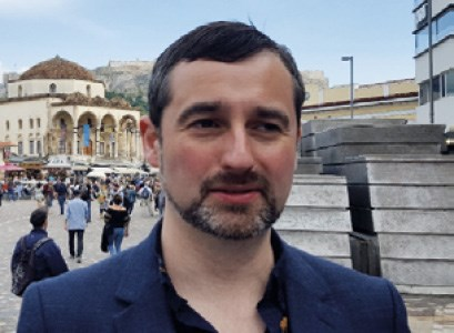 A man with short dark hair and a beard standing in a bustling outdoor area with historical architecture and people in the background.