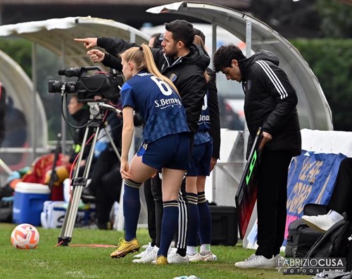Football players and coaches on the sideline during a match, with one player wearing a jersey with the number 6, and several team staff members giving instructions beside a substitution board.