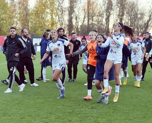 A group of female football players wearing white jerseys celebrating on a grassy field, with some teammates and coaches joining in the background. Trees line the rear of the field.