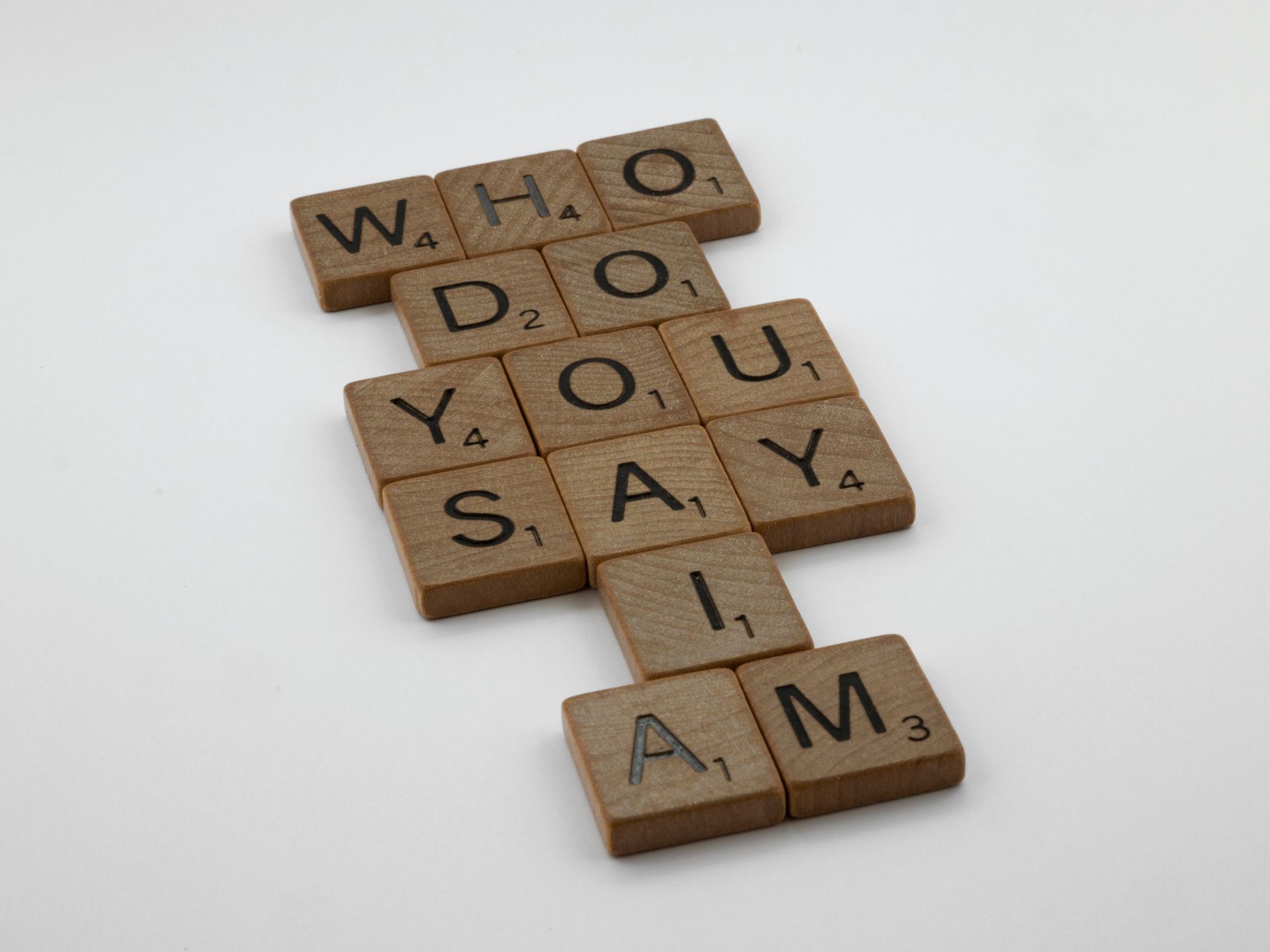Wooden Scrabble tiles arranged to spell the words "WHO DO YOU SAY I AM" on a white surface.