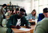 Effective peer feedback Students seated at desks in a classroom, taking an exam or test, with a teacher walking around in the background.