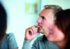 Listening as a manager A thoughtful man with grey hair, wearing a grey sweater, sitting in profile view with a finger resting on his lips, in a contemplative pose. Blurred people are visible in the foreground, indicating a group setting.