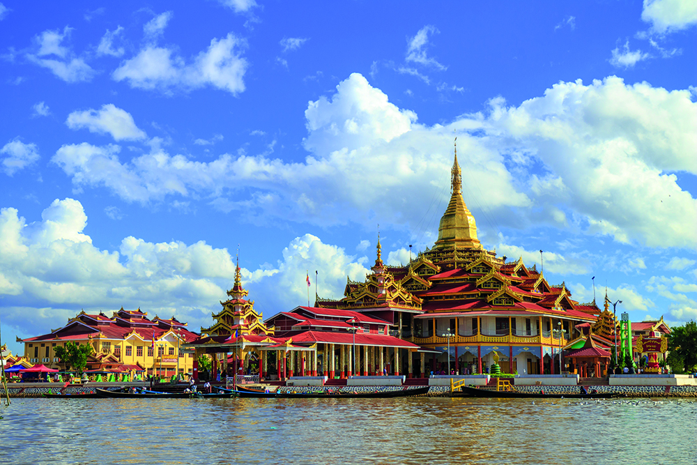 A colourful pagoda with a golden spire sits by a river, surrounded by traditional red and yellow roofs under a blue sky with scattered clouds.