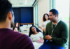 SLT and CLL in the ELT classroom: revisited A man wearing a green sweater and an ID lanyard is speaking to a group of attentive students in a brightly lit classroom. The students are seated around him, listening and engaging in the discussion.